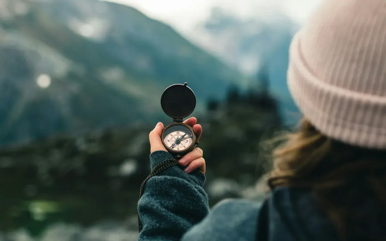 A person in a pink beanie holds a compass, navigating a misty mountain landscape. This symbolizes the journey of transition coaching.