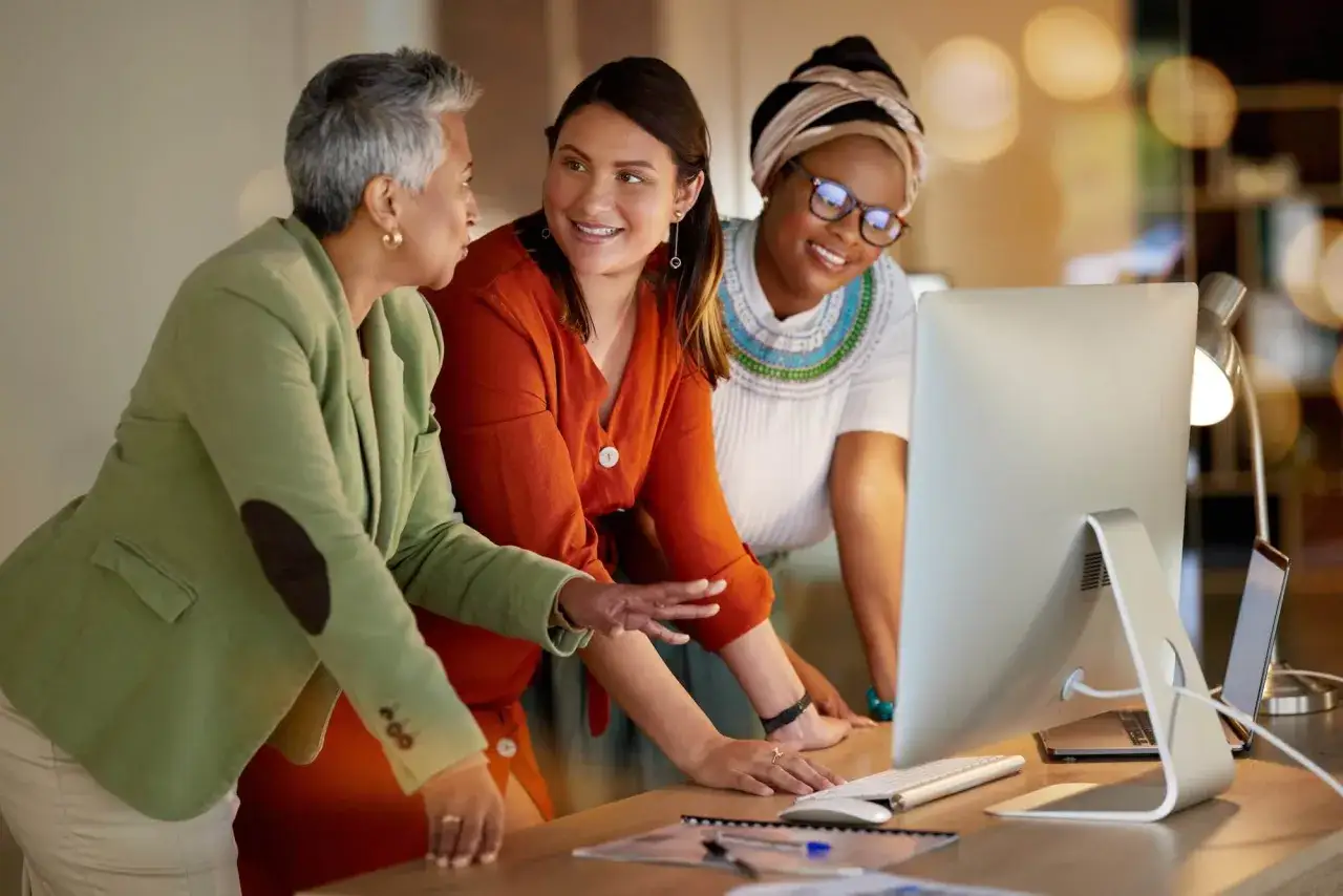 Three women collaborate around a computer, discussing ideas. This scene illustrates why coaching is essential for team growth and problem-solving.